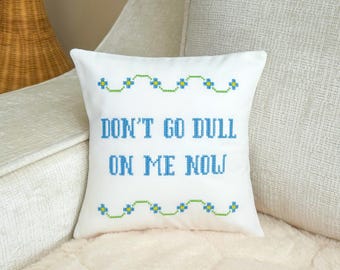 May include: A square white throw pillow featuring blue cross-stitch lettering that reads "DON'T GO DULL ON ME NOW." The text is framed by two horizontal rows of green and blue floral cross-stitch patterns at the top and bottom of the pillow.