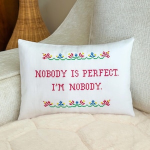 May include: A rectangular white throw pillow featuring a cross-stitch design. The text reads NOBODY IS PERFECT. I'M NOBODY. in red letters, flanked by decorative floral borders in red, blue, yellow, and green cross-stitch embroidery patterns.