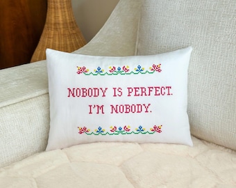 May include: A rectangular white throw pillow featuring a cross-stitch design. The text reads NOBODY IS PERFECT. I'M NOBODY. in red letters, flanked by decorative floral borders in red, blue, yellow, and green cross-stitch embroidery patterns.