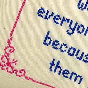 May include: A cream-colored fabric piece featuring blue cross-stitched text that reads "We everyone because them". A decorative pink cross-stitched border frames the left side of the fabric, creating a distinct corner pattern with intricate geometric details.