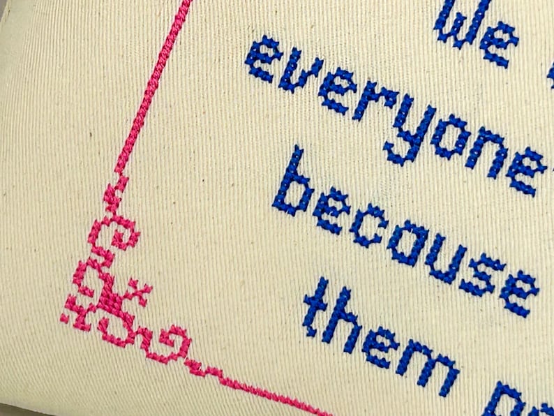 May include: A cream-colored fabric piece featuring blue cross-stitched text that reads "We everyone because them". A decorative pink cross-stitched border frames the left side of the fabric, creating a distinct corner pattern with intricate geometric details.