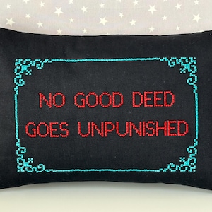 May include: A rectangular black throw pillow featuring cross-stitched embroidery. The text "NO GOOD DEED GOES UNPUNISHED" is stitched in red thread in the center, surrounded by a decorative teal border with ornate corner flourishes.