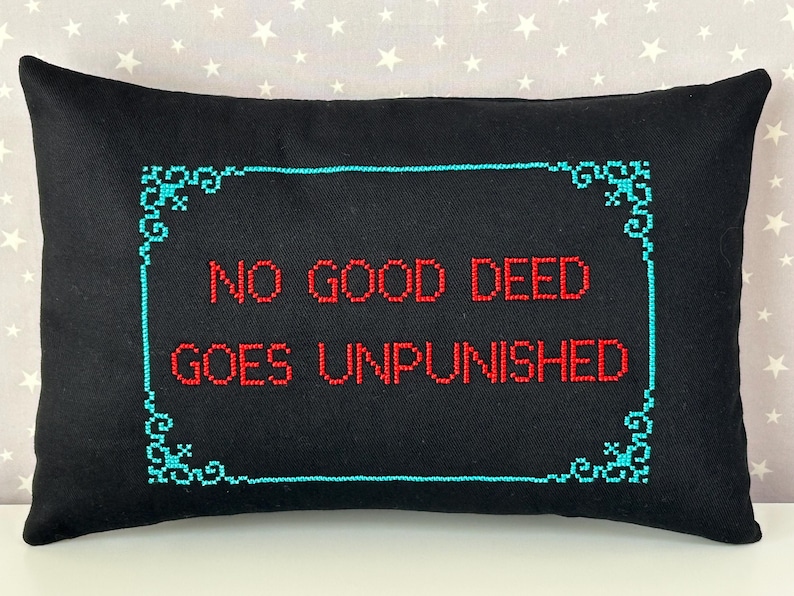 May include: A rectangular black throw pillow featuring cross-stitched embroidery. The text "NO GOOD DEED GOES UNPUNISHED" is stitched in red thread in the center, surrounded by a decorative teal border with ornate corner flourishes.