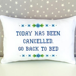 Puede incluir: Almohada blanca con bordado azul que dice "Hoy ha sido cancelado. Vuelve a la cama." El bordado está rodeado de diseños florales verdes y azules.