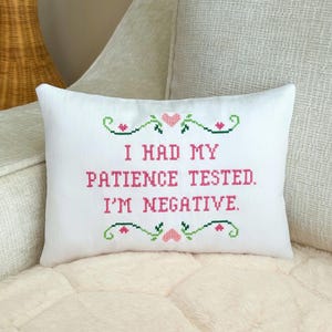 May include: A rectangular white throw pillow featuring a cross-stitch design with the text I HAD MY PATIENCE TESTED. I'M NEGATIVE. in pink letters, accented by green decorative scrollwork and small pink hearts above and below the text.