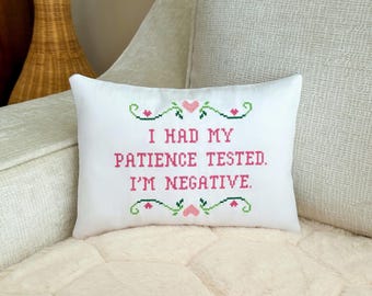 May include: A rectangular white throw pillow featuring a cross-stitch design with the text I HAD MY PATIENCE TESTED. I'M NEGATIVE. in pink letters, accented by green decorative scrollwork and small pink hearts above and below the text.