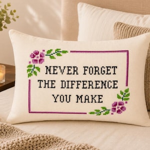 May include: A rectangular off-white throw pillow featuring a cross-stitch design with a purple border. The text reads NEVER FORGET THE DIFFERENCE YOU MAKE in black letters, accented by purple flowers and green leaves at the corners of the frame.