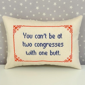 May include: A rectangular off-white throw pillow featuring a cross-stitched design. The text reads, "You can't be at two congresses with one butt." The lettering is in dark blue, enclosed within a decorative orange rectangular border.