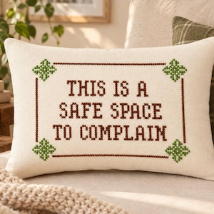 May include: A rectangular off-white throw pillow featuring a cross-stitch design with the brown text "THIS IS A SAFE SPACE TO COMPLAIN" enclosed in a brown rectangular border with green cross-stitch floral accents at each of the four corners.