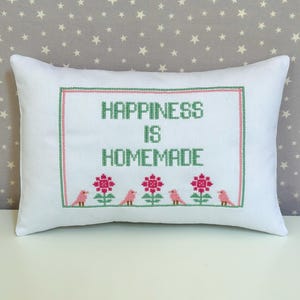 May include: White rectangular pillow with cross-stitch embroidery. The pillow features the text "HAPPINESS IS HOMEMADE" in green, framed by a green and pink border. Below the text are pink flowers and birds.