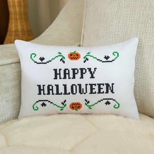 May include: A rectangular white throw pillow featuring black cross-stitch embroidery that reads HAPPY HALLOWEEN. The text is framed by green vine patterns, small black hearts, and two orange jack-o'-lantern pumpkin motifs.