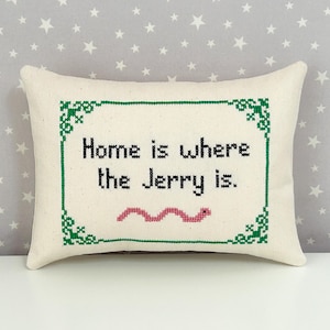 May include: A rectangular off-white throw pillow featuring a cross-stitched design with a green decorative border. The black text reads "Home is where the Jerry is." Below the text is a small, pink, wavy, cross-stitched worm illustration.