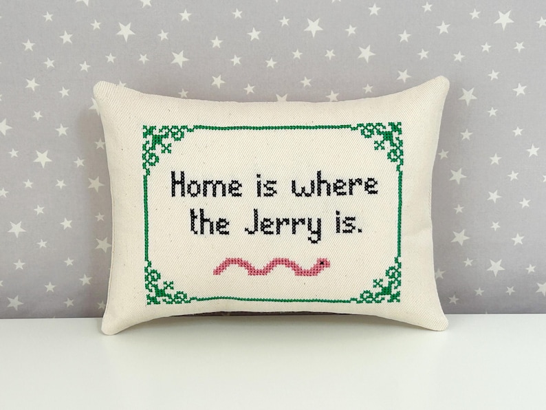 May include: A rectangular off-white throw pillow featuring a cross-stitched design with a green decorative border. The black text reads "Home is where the Jerry is." Below the text is a small, pink, wavy, cross-stitched worm illustration.
