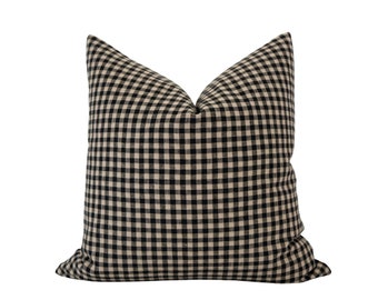 May include: Square throw pillow with a black and beige gingham pattern. The pillow has a soft, textured appearance and is designed for decorative use on a bed or sofa. The pillow is square-shaped.