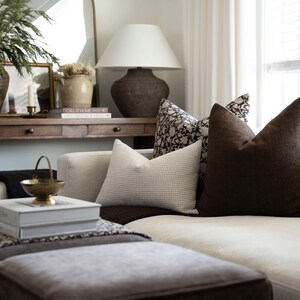 Op de afbeelding: Een witte bank met bruine kussens en een kussen met een patroon. De bank staat in een woonkamer met een houten consoletafel erachter. De consoletafel heeft een lamp, boeken en een vaas met gedroogde bloemen.