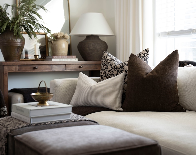 Op de afbeelding: Een witte bank met bruine kussens en een kussen met een patroon. De bank staat in een woonkamer met een houten consoletafel erachter. De consoletafel heeft een lamp, boeken en een vaas met gedroogde bloemen.