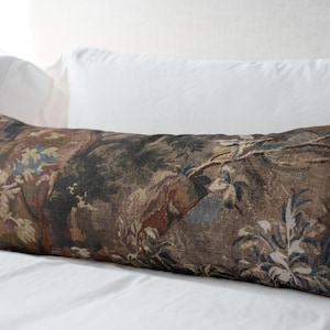 Puede incluir: Un coj&iacute;n decorativo rectangular con un dise&ntilde;o de tapiz que representa una escena de paisaje en tonos marrones, verdes y azules. El coj&iacute;n est&aacute; colocado sobre una cama blanca con dos almohadas blancas detr&aacute;s.
