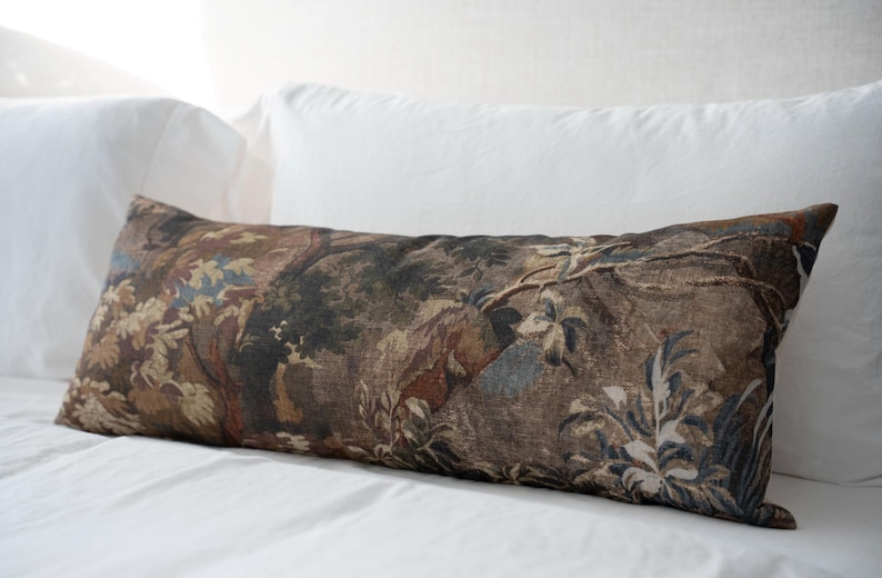 Puede incluir: Un coj&iacute;n decorativo rectangular con un dise&ntilde;o de tapiz que representa una escena de paisaje en tonos marrones, verdes y azules. El coj&iacute;n est&aacute; colocado sobre una cama blanca con dos almohadas blancas detr&aacute;s.