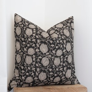 May include: Square throw pillow with a black background and a repeating floral pattern in a light beige color. The pillow is placed on a wooden surface, with a white curtain in the background. The pillow has a soft, textured appearance.