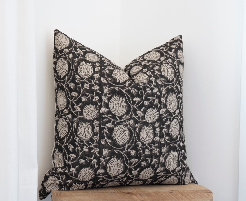 May include: Square throw pillow with a black background and a repeating floral pattern in a light beige color. The pillow is placed on a wooden surface, with a white curtain in the background. The pillow has a soft, textured appearance.
