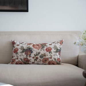 May include: A floral patterned throw pillow with a beige background and red, orange, and green flowers. The pillow is on a beige sofa.
