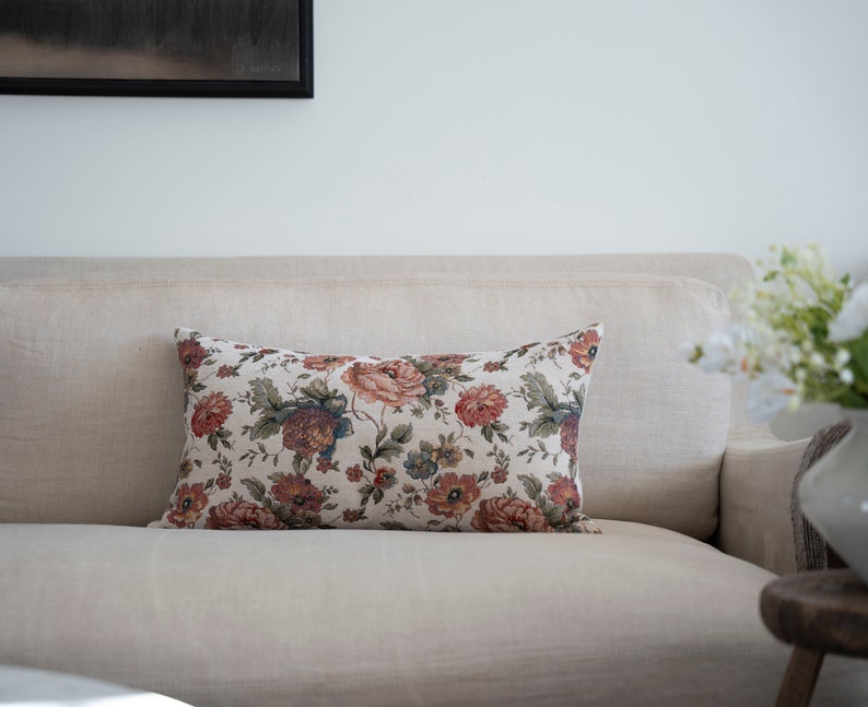 May include: A floral patterned throw pillow with a beige background and red, orange, and green flowers. The pillow is on a beige sofa.