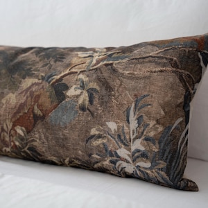 Puede incluir: Un coj&iacute;n rectangular decorativo con un dise&ntilde;o de tapiz. El coj&iacute;n presenta un patr&oacute;n detallado de una escena de bosque con &aacute;rboles, follaje y toques de tonos azules y marrones. El coj&iacute;n est&aacute; sobre una superficie blanca.