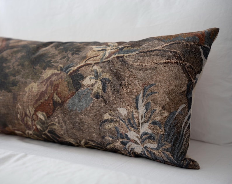 Puede incluir: Un coj&iacute;n rectangular decorativo con un dise&ntilde;o de tapiz. El coj&iacute;n presenta un patr&oacute;n detallado de una escena de bosque con &aacute;rboles, follaje y toques de tonos azules y marrones. El coj&iacute;n est&aacute; sobre una superficie blanca.