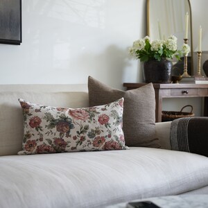 May include: A white linen sofa with a floral patterned cushion and a brown striped cushion. A brown throw blanket is draped over the arm of the sofa.