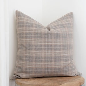 May include: A brown and beige plaid throw pillow with a square shape. The pillow is sitting on a wooden surface.