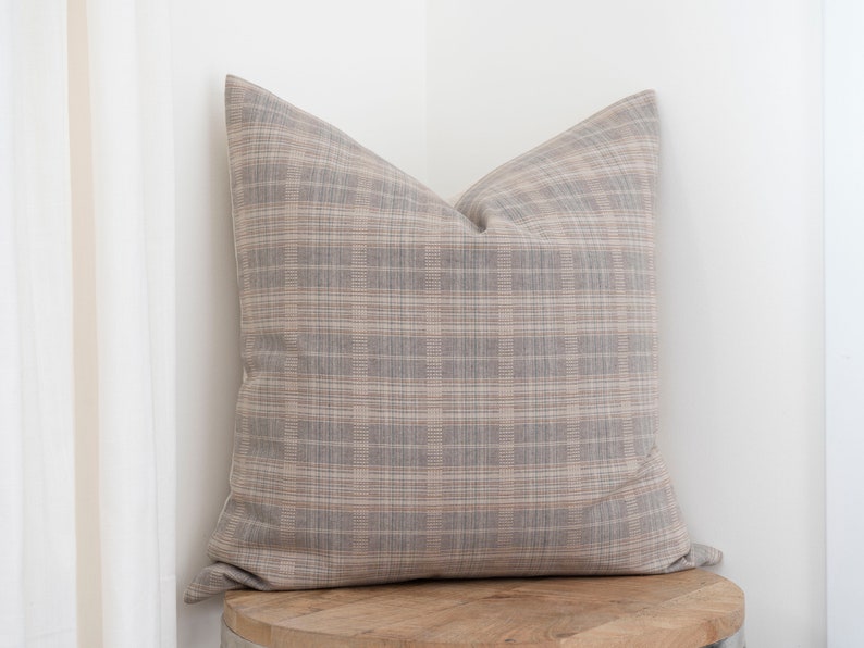 May include: A brown and beige plaid throw pillow with a square shape. The pillow is sitting on a wooden surface.