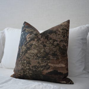 May include: A decorative throw pillow with a detailed landscape design in shades of brown, green, and blue. The pillow features a floral and tree pattern, creating a vintage aesthetic. The pillow is placed on a white bed.