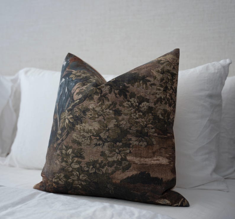May include: A decorative throw pillow with a detailed landscape design in shades of brown, green, and blue. The pillow features a floral and tree pattern, creating a vintage aesthetic. The pillow is placed on a white bed.