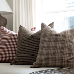May include: Three decorative throw pillows on a white couch. The pillows are made of a brown and white plaid fabric, a brown and white striped fabric, and a red and white gingham fabric.