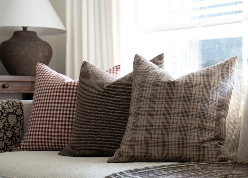 May include: Three decorative throw pillows on a white couch. The pillows are made of a brown and white plaid fabric, a brown and white striped fabric, and a red and white gingham fabric.