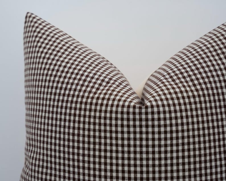 K&ouml;nnte beinhalten: Nahaufnahme eines kissens mit braun-wei&szlig;em Vichy-Muster. Das Kissen hat ein klassisches Karomuster mit hellem Hintergrund und dunkelbraunen Quadraten. Es ist ein dekorativer Artikel f&uuml;r ein Bett oder Sofa.