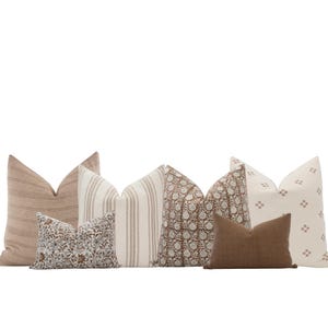 Könnte beinhalten: Eine Sammlung dekorativer Zierkissen in verschiedenen Mustern und Farben. Die Kissen umfassen gestreifte, florale und unifarbene Designs in neutralen Tönen von Beige, Braun und Creme. Die Anordnung deutet auf ein Wohnkulturthema hin.