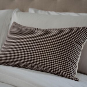 K&ouml;nnte beinhalten: Ein rechteckiges Kissen mit einem braun-wei&szlig;en Vichy-Muster. Das Kissen liegt auf einem wei&szlig;en Bett mit anderen wei&szlig;en Kissen. Das Kissen hat eine neutrale Farbpalette und ein klassisches Design.