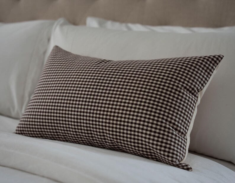 K&ouml;nnte beinhalten: Ein rechteckiges Kissen mit einem braun-wei&szlig;en Vichy-Muster. Das Kissen liegt auf einem wei&szlig;en Bett mit anderen wei&szlig;en Kissen. Das Kissen hat eine neutrale Farbpalette und ein klassisches Design.