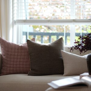 May include: A white couch with three pillows. The pillows are red and white gingham, brown and white striped, and beige and white gingham. A book is on the couch in front of the pillows.