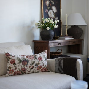 May include: A white sofa with a floral patterned cushion, a brown throw blanket, and a wooden console table with a vase of white flowers, a lamp, and candles.