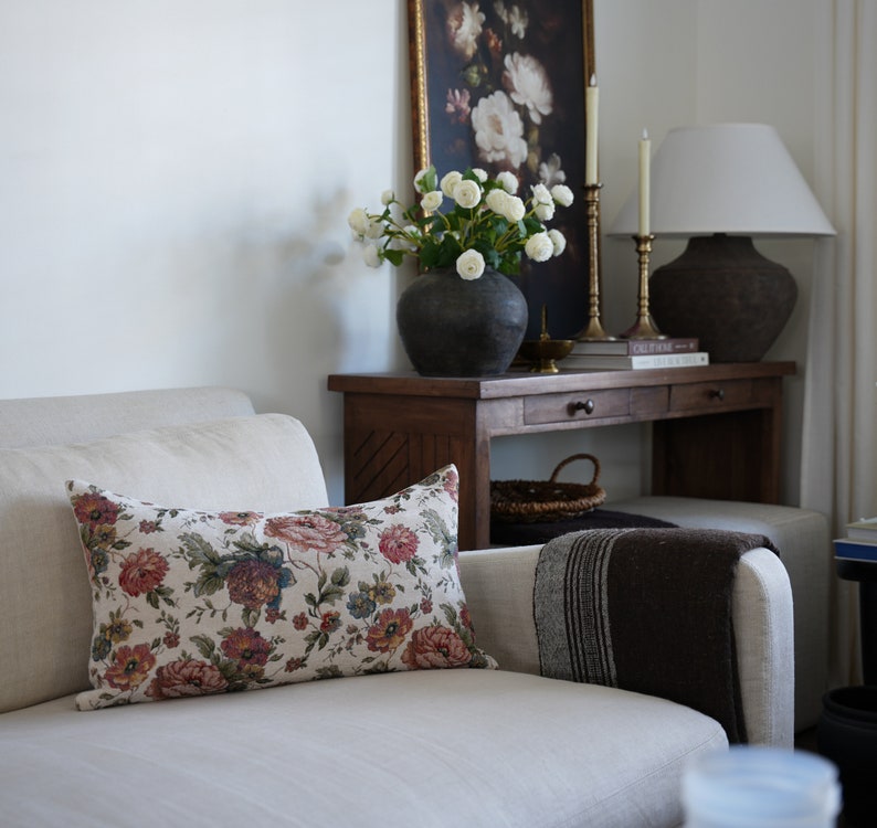 May include: A white sofa with a floral patterned cushion, a brown throw blanket, and a wooden console table with a vase of white flowers, a lamp, and candles.