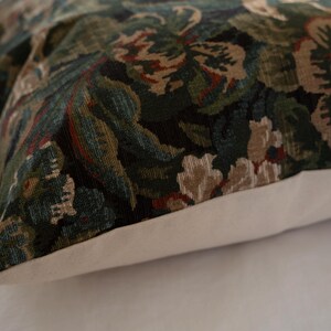 May include: Decorative pillow with a floral pattern in shades of green, brown, and cream. The pillow has a white border and a textured fabric. The design features stylized flowers and foliage.