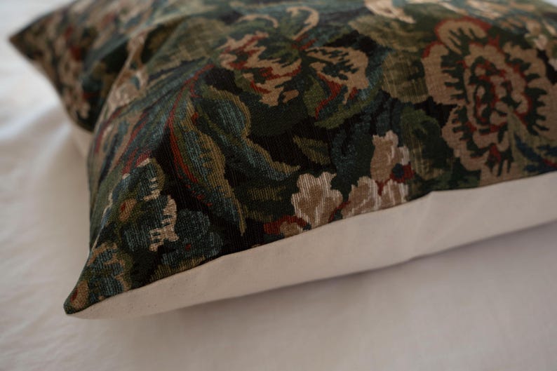 May include: Decorative pillow with a floral pattern in shades of green, brown, and cream. The pillow has a white border and a textured fabric. The design features stylized flowers and foliage.