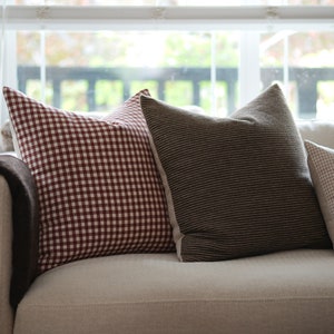 May include: A white couch with three throw pillows. The pillows are red and white gingham, brown and white striped, and beige and white gingham.