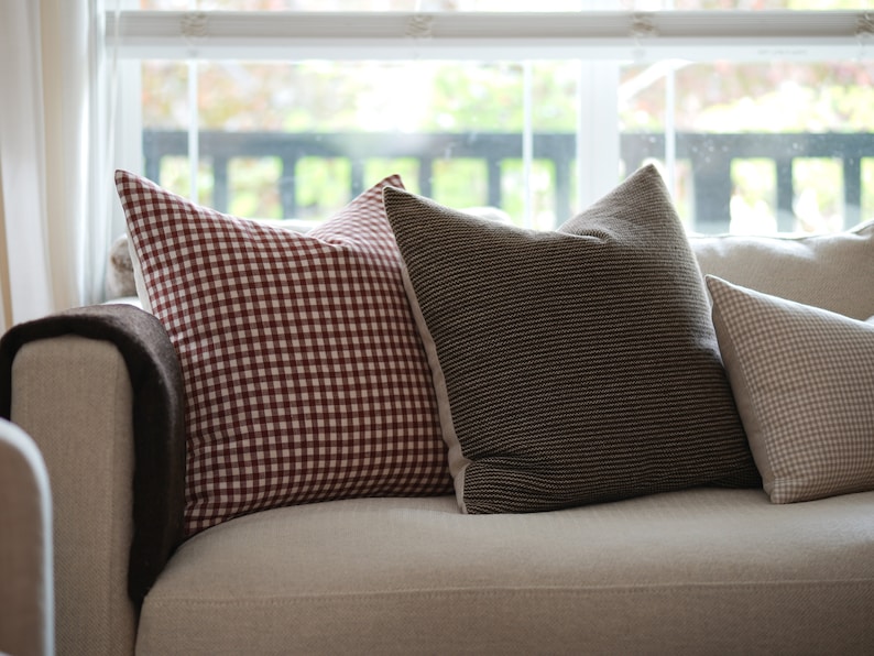 May include: A white couch with three throw pillows. The pillows are red and white gingham, brown and white striped, and beige and white gingham.