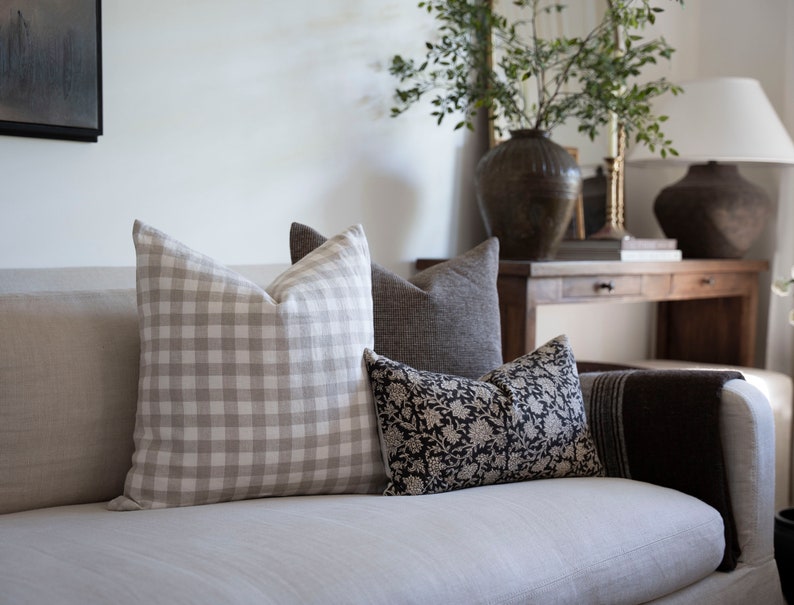 Op de afbeelding: Een witte bank met een aantal kussens. De kussens zijn in een beige en wit ruitpatroon, een donkergrijs linnen en een zwart-wit bloemenpatroon. De bank staat in een woonkamer met een houten tafel en een vaas met groen in de achtergrond.
