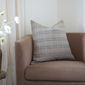 May include: A brown armchair with a plaid throw pillow. The armchair is in a room with white curtains and a vase of white flowers.