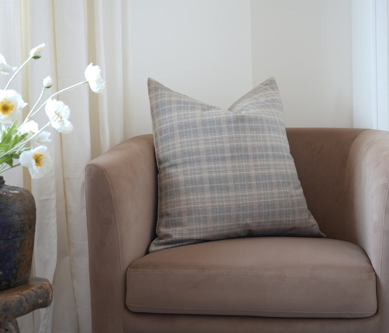 May include: A brown armchair with a plaid throw pillow. The armchair is in a room with white curtains and a vase of white flowers.