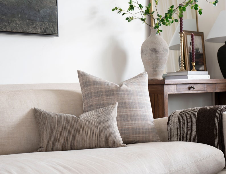 May include: A white linen sofa with two throw pillows, one with a plaid pattern and one with a striped pattern. A brown throw blanket is draped over the back of the sofa.